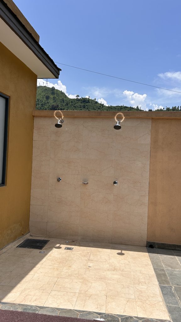 An outdoor shower area with three shower heads mounted on a beige tiled wall. Background contains blue clear sky with some clouds and green hills.