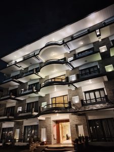 A modern multi-story building with a textured stone facade and multiple balconies, illuminated by soft lighting at night. 