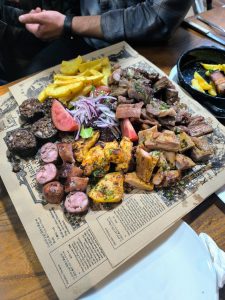 A large platter of assorted meats is presented on a decorative paper, featuring various types of grilled meats including sausages, chicken, and beef, alongside sides of fried plantains, fresh tomato slices, and red onions