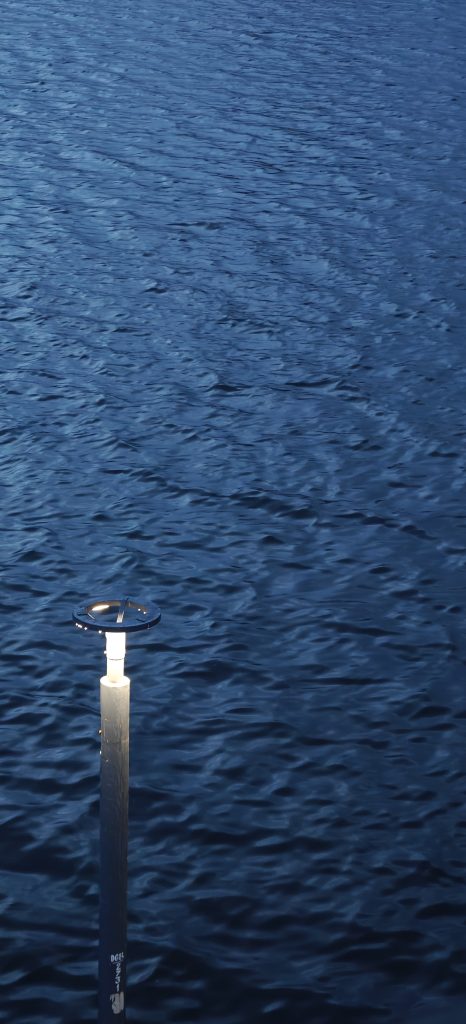 A solitary lamp post standing beside calm, deep blue water, with soft ripples across the surface.