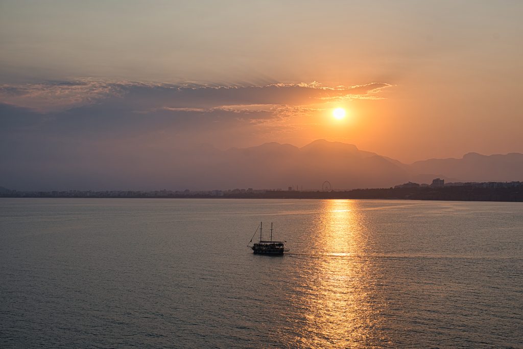 Sunset over the sea with a sailboat, golden light reflecting on the water, and mountains in the background.