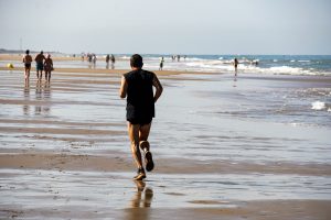 A person jogging along a wide, sandy beach, with the ocean in the background. Other beachgoers can be seen walking at a distance, and the water reflects the sunlight, creating a shimmering effect on the wet sand.