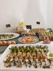 The image platter of Sushi arranged in different shapes of plates like rectangular, circle. The picture contains veg sushi, salmon sushi, nigiri sushi with a bowl of sauce in between the plates.