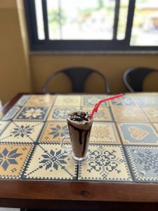 A tall glass of chocolate milkshake topped with whipped cream and chocolate drizzle sits on a patterned table. 