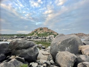 Calm waters of the Tungabhadra River reflect the sky, with rocks and a hill showcasing Hampi’s natural beauty.