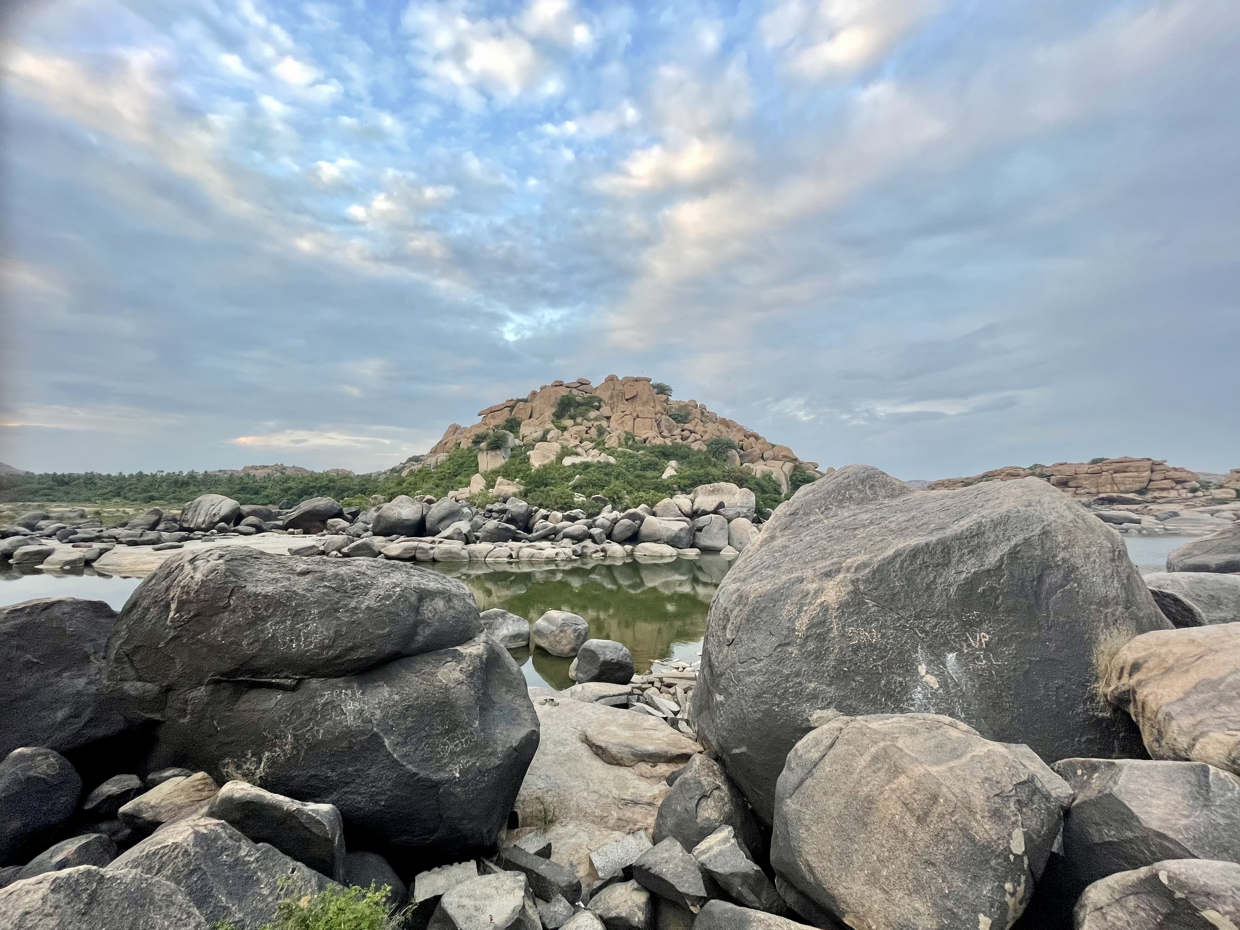 Calm waters of the Tungabhadra River reflect the sky, with rocks and a hill showcasing Hampi’s natural beauty.