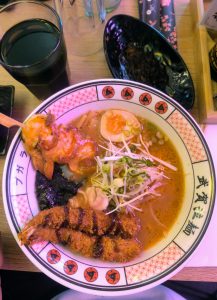 A bowl of ramen with rich, golden broth is presented on a colorful dish. Toppings include sliced soft-boiled egg, green onions, and a piece of nori seaweed. 