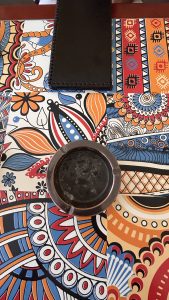 A colorful tablecloth featuring intricate floral and geometric patterns in vibrant colors such as orange, blue, black, and red. In the center, there is a round, dark-colored ashtray with a smooth finish.