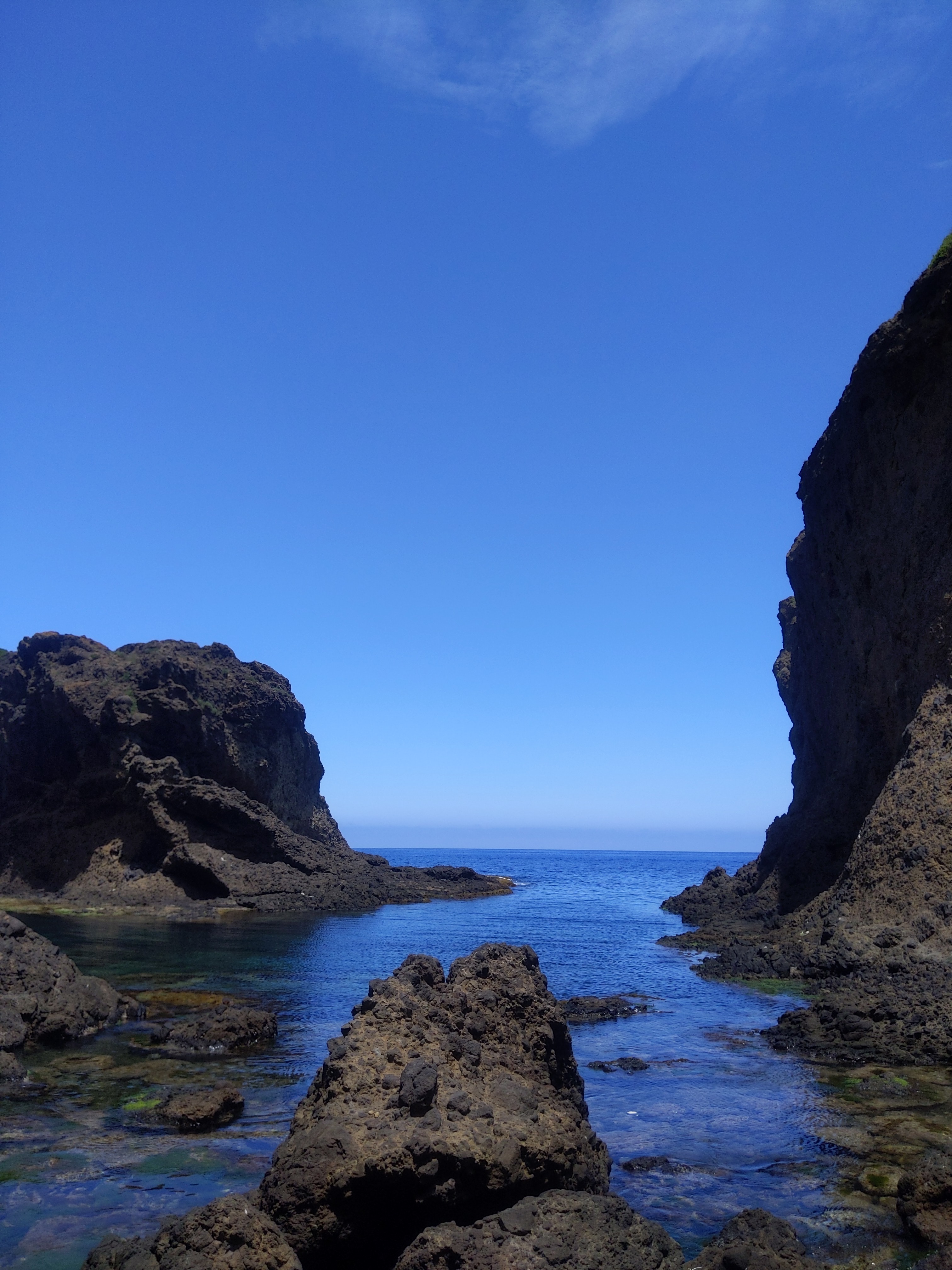 A cove with dark rock formations on either side, framing a bright blue ocean under a clear sky.
