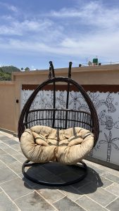 A cozy, suspended hanging chair with a circular frame and soft beige cushions sits on a tiled floor. Behind the chair is a mural featuring stylized drawings of plants and animals on a white wall, adding an artistic backdrop. The sky above is bright blue with a few wispy clouds, indicating a sunny day.