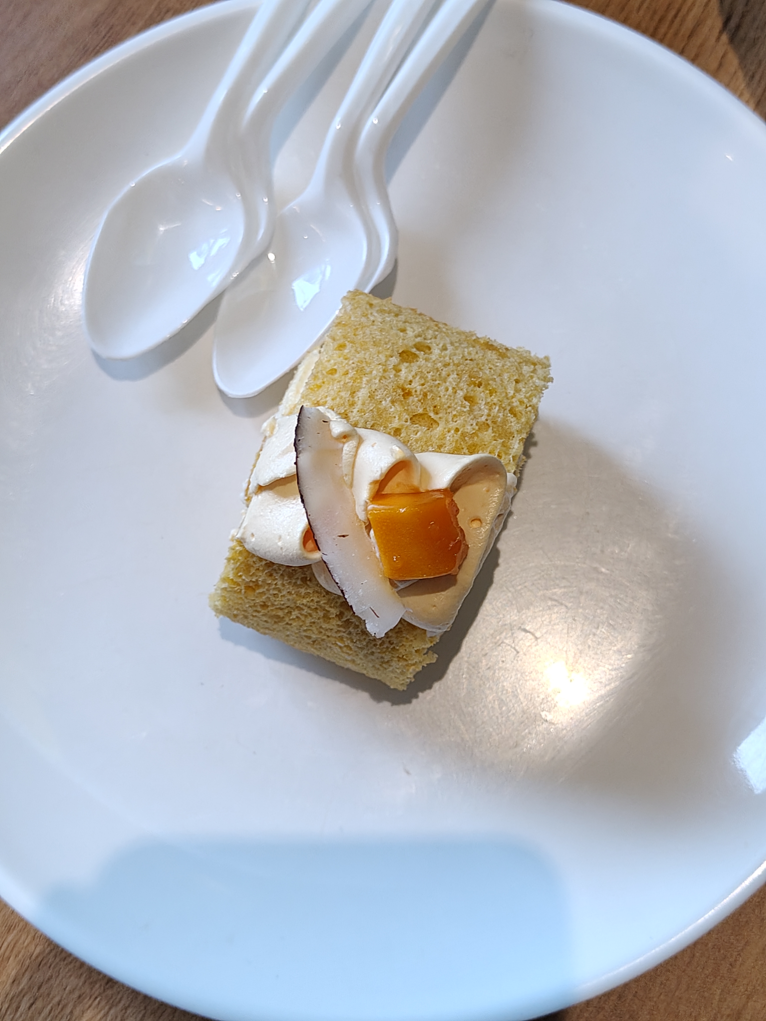 A small piece of yellow sponge cake topped with a dollop of cream, adorned with slices of coconut and a piece of orange jelly, sits on a white plate.