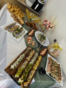 A picture of a table containing different varietes of foods which includes samosa, sel roti(famous nepali dish), drinks and mocktails, bread rolls, nuts wrapped in a plastic, a vase with beautiful flowers, different varieties of sushi served on different shapes and sizes of plates.