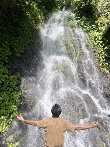 A person stands with their arms outstretched in front of a cascading waterfall surrounded by lush green foliage. 