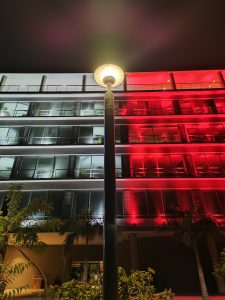 Night-light captures a striking contrast of warm streetlamp glow against a modern building lit in white and red tones. The centered pole divides the frame symmetrically, adding depth and balance. A blend of architecture, color, and calm nighttime ambiance.