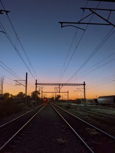 A railway track stretches into the distance at dusk, flanked by electrical poles and wires. The sky transitions from deep blue to a warm orange and purple hue as the sun sets