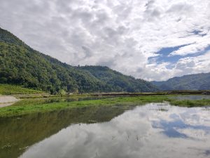 A serene landscape featuring a calm waterland surrounded by gentle hills, with lush green trees dotting the slopes and reflecting softly on the water&rsquo;s surface. A sky covered with clouds. 