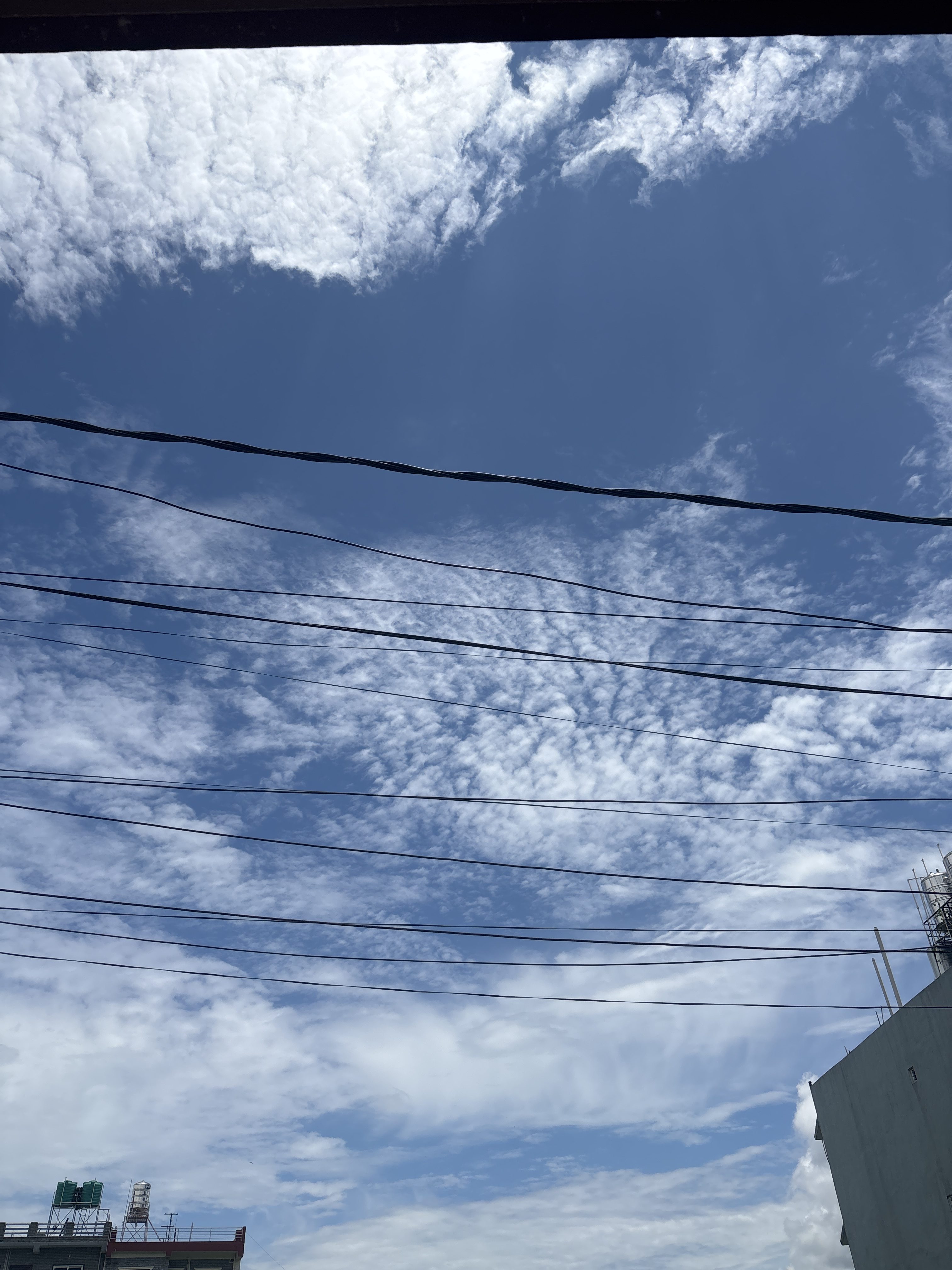 A clear blue sky partially covered with fluffy white clouds, intersected by several black power lines.