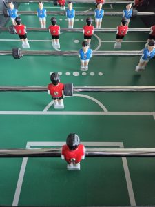 A foosball table with red and blue plastic players on metal rods, set on a green field marked with white pitch lines.