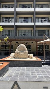 A landscaped area featuring a large, smooth stone sculpture in the foreground, accompanied by smaller stones.