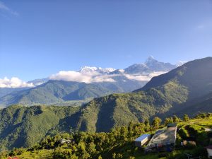 A panoramic view of lush green mountains under a clear blue sky, with snow-capped peaks in the distance partially obscured by clouds.