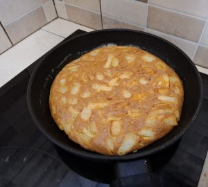 A golden-brown tortilla-like dish sits in a black frying pan on a stove. 