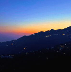A picturesque twilight scene featuring a mountainous silhouette against a gradient sky transitioning from deep blue at the top to warm orange and purple hues near the horizon.
