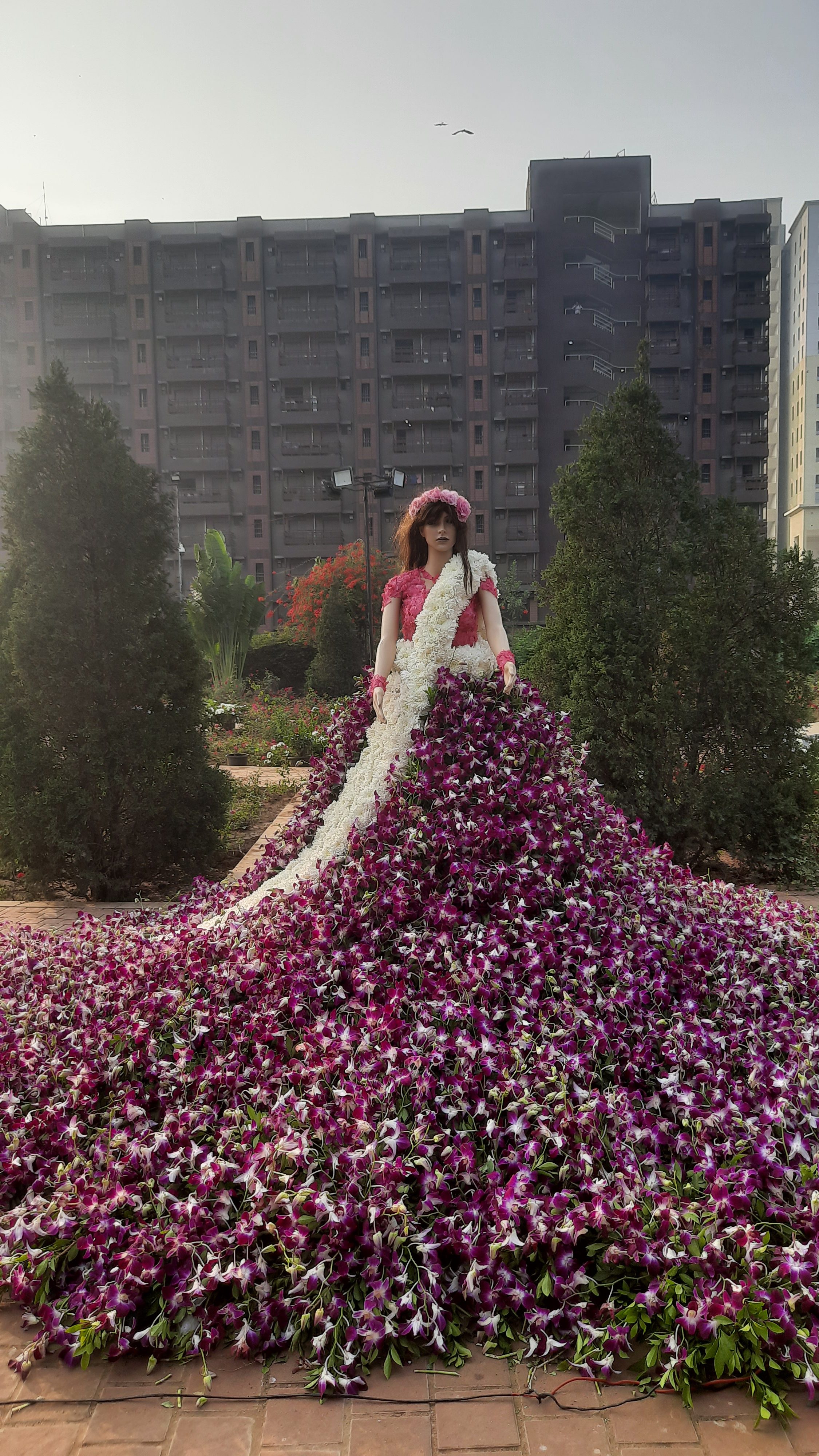 An image containing a doll of flower. The doll is dress with colourful flowers from head to toe. It is in the garden in front of a building.