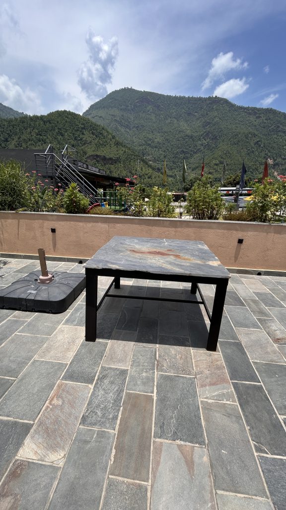 A black stone table is positioned on a patterned stone patio under a partly cloudy sky. A clear view of green hills, flowers and flags on the background.