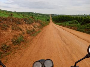 View from a motorbike in dirt road that stretches into the distance, flanked by lush green fields and hills. 