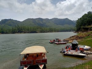 Boats docked along a calm lake surrounded by lush green hills and pine trees in Kerala, India, with tourists boarding for a ride under a partly cloudy sky.
