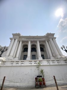 A low-angle view of a large, neoclassical building with white columns and intricate architectural details.
