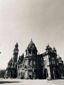 The monochromatic photo showcases the grand, ornate Indo-Saracenic architecture of the New Palace in Kolhapur, Maharashtra. The building features multiple domes, arches, and a prominent clock tower on the left. Its intricate details are highlighted by the stark black and white tones, set against a bright, clear sky.
