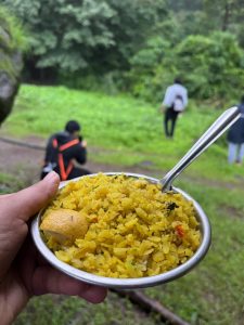 Poha served in a steel plate during a mountain trek. Closeup shot of yellow rice in a metal bowl with a spoon sticking out. People in the background in a forest setting.