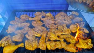A grill filled with numerous pieces of marinated chicken, colored in a golden hue, cooking over an open flame. Smoke rises around the chicken as it sizzles.