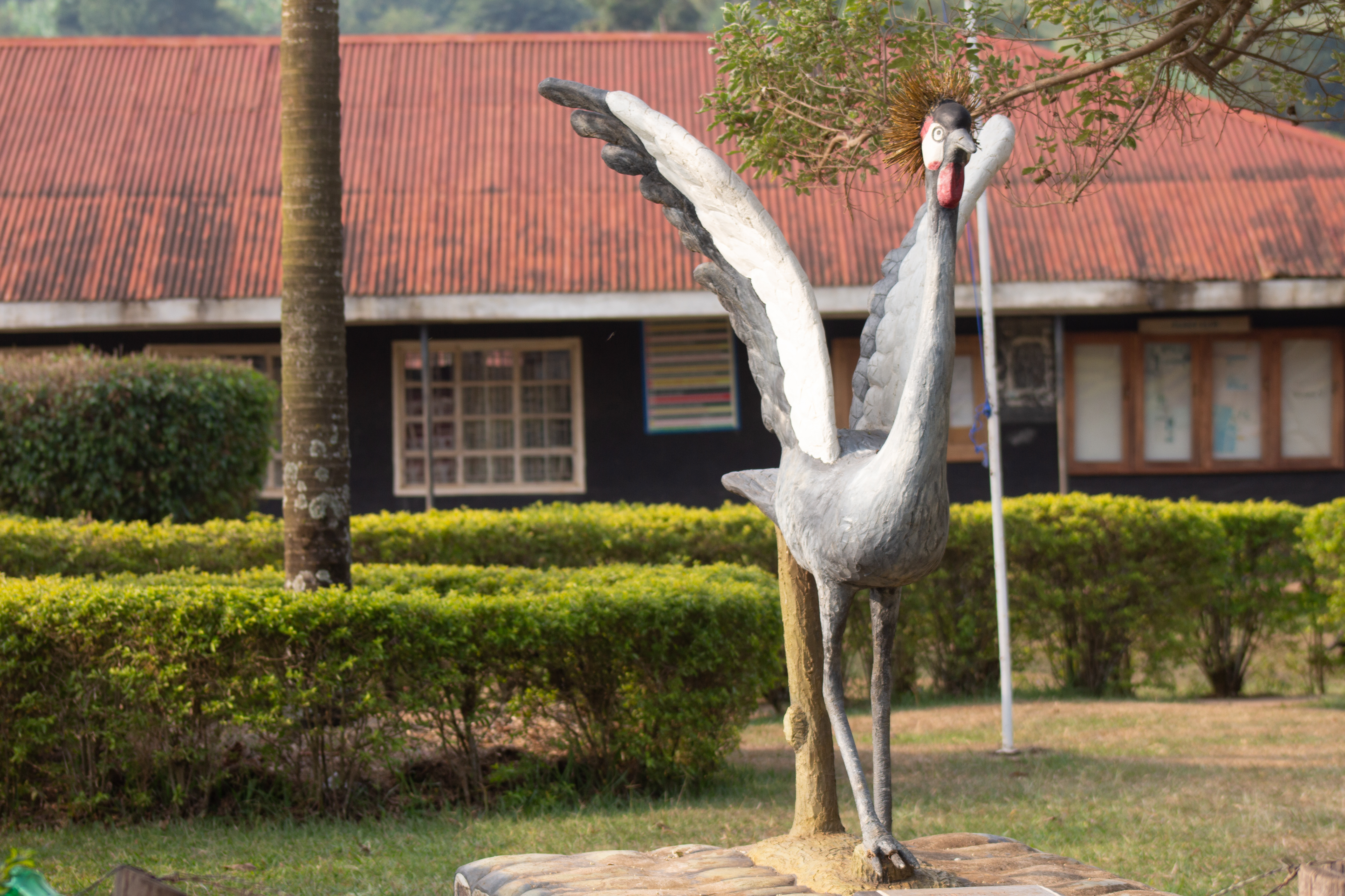 A decorative sculpture of a crane stands in a garden area, with vibrant greenery and neatly trimmed bushes surrounding it.