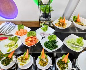 A vibrant display of fresh vegetables and salads arranged in white bowls. The assortment includes sliced cucumbers, tomatoes, carrots, onions, green chilies, and lime, all garnished with leafy lettuce.