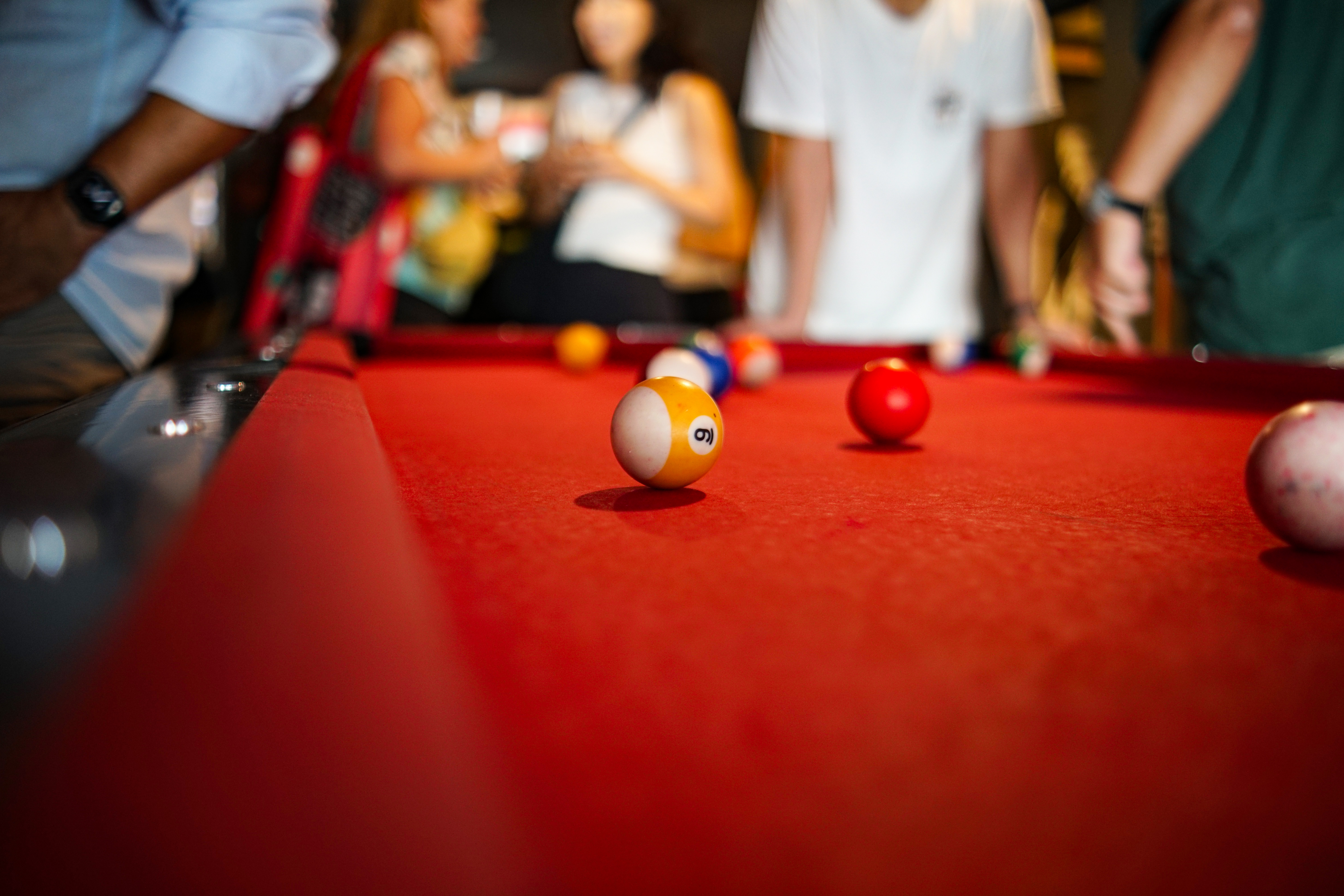 A close-up view of a billiards table with a red felt surface, featuring several colorful pool balls scattered across it, including a prominent yellow ball with the number 9. In the background, people are casually interacting