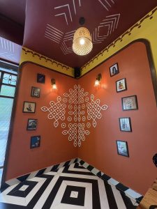 An image of interior of a restaurant whose wall is painted with unique art and photo frames representing the historical art and culture. A beautiful lamp is hung on the ceiling. 