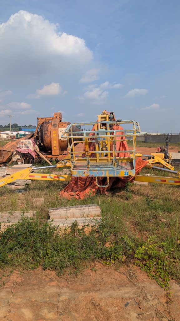 A construction site featuring heavy machinery, including a large concrete mixer partially covered with a tarp and a yellow elevated work platform.
