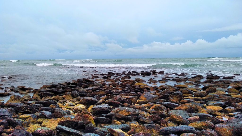 A rocky sea shore with waves crashing against the stones under a cloudy sky.