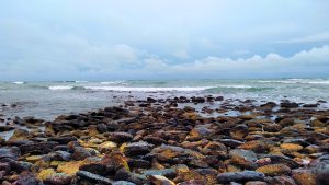 A rocky sea shore with waves crashing against the stones under a cloudy sky.