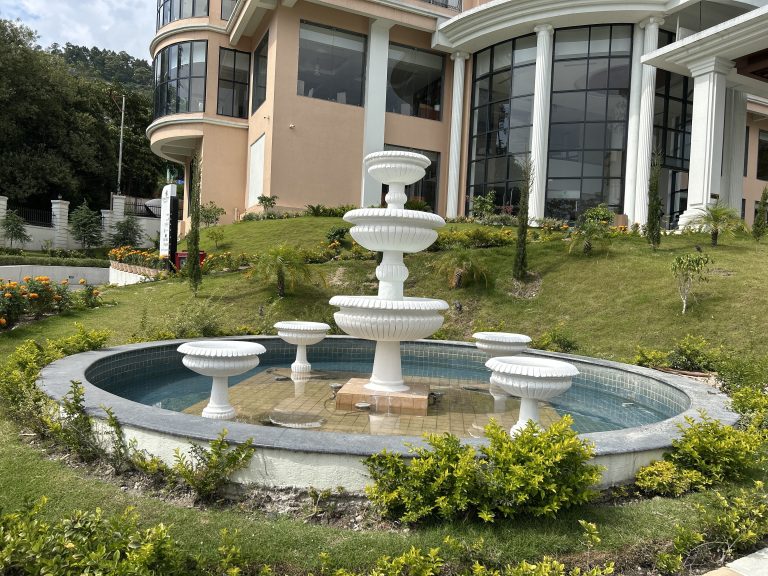 The image features a tiered white fountain situated in a circular pool, surrounded by greenery and decorative plants