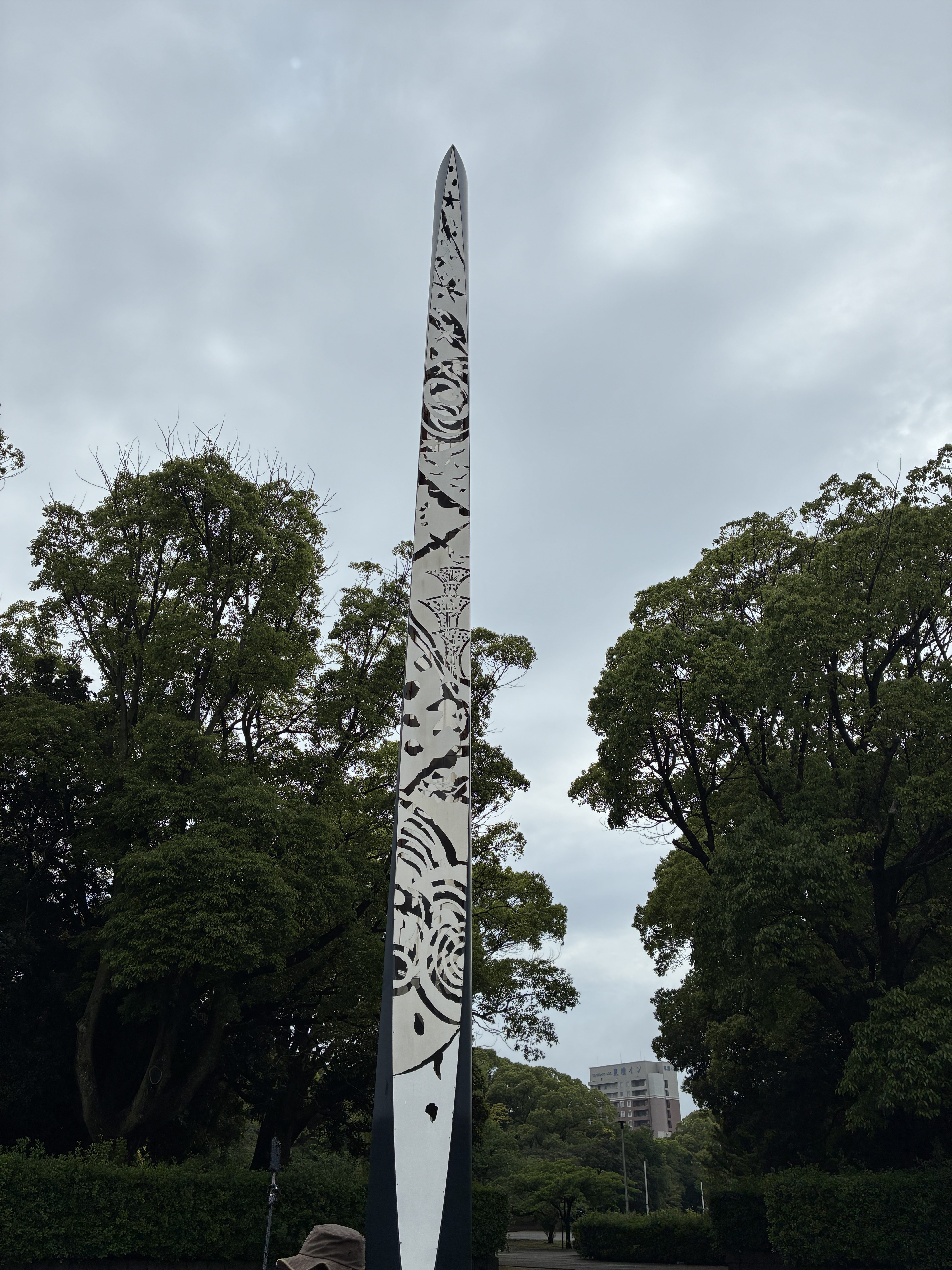 
A tall, slender metallic sculpture with intricate etchings stands in Chiba Port Park, surrounded by lush green trees.