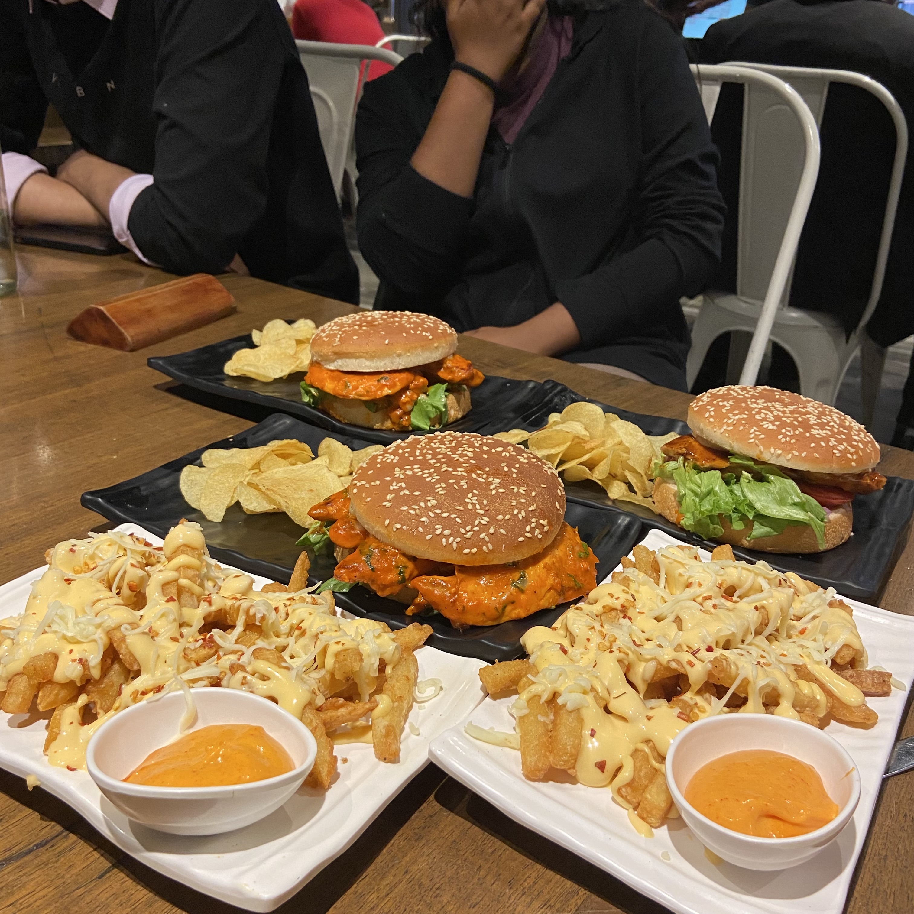 A wooden table holds two plates with sesame seed burgers—one with chicken, the other with lettuce and tomato—plus crispy chips, cheesy fries with chili flakes, orange dipping sauce, and a pizza.