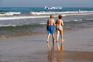 An elder couple walks hand in hand along a sandy beach, their feet touching the water's edge. The woman is wearing a colorful swimsuit, while the man is dressed in blue swim shorts and a hat.