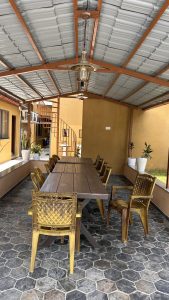A shaded outdoor dining area featuring a long wooden table surrounded by several decorative chairs.