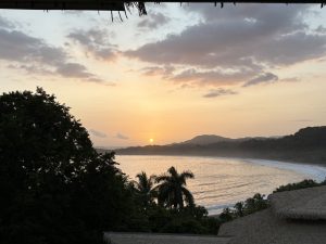 Atardecer Playa Carrillo, Costa Rica at dusk, with the setting sun reflecting orange on the water, and the beach curved around the sea. There is the thatched roof of a building and trees in the foreground and hills in the background.