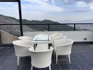 A rooftop terrace featuring a glass-top dining table surrounded by seven white wicker chairs.

