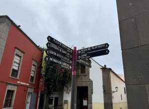 Tourist signage in the foreground, with arrows pointing to Plaza de Santa Ana, the Cathedral of Santa Ana, the Center for Plastic Arts, the CAAM, Plaza del Pilar Nuevo, the Tourist Information, and the Vegueta Market. In the background, the colonial facades of San Antonio Abad street can be seen, including the small adjacent chapel.
