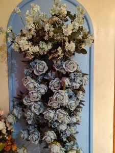 A decorative arrangement of artificial flowers, prominently featuring light blue roses, surrounded by clusters of smaller flowers and greenery. 
